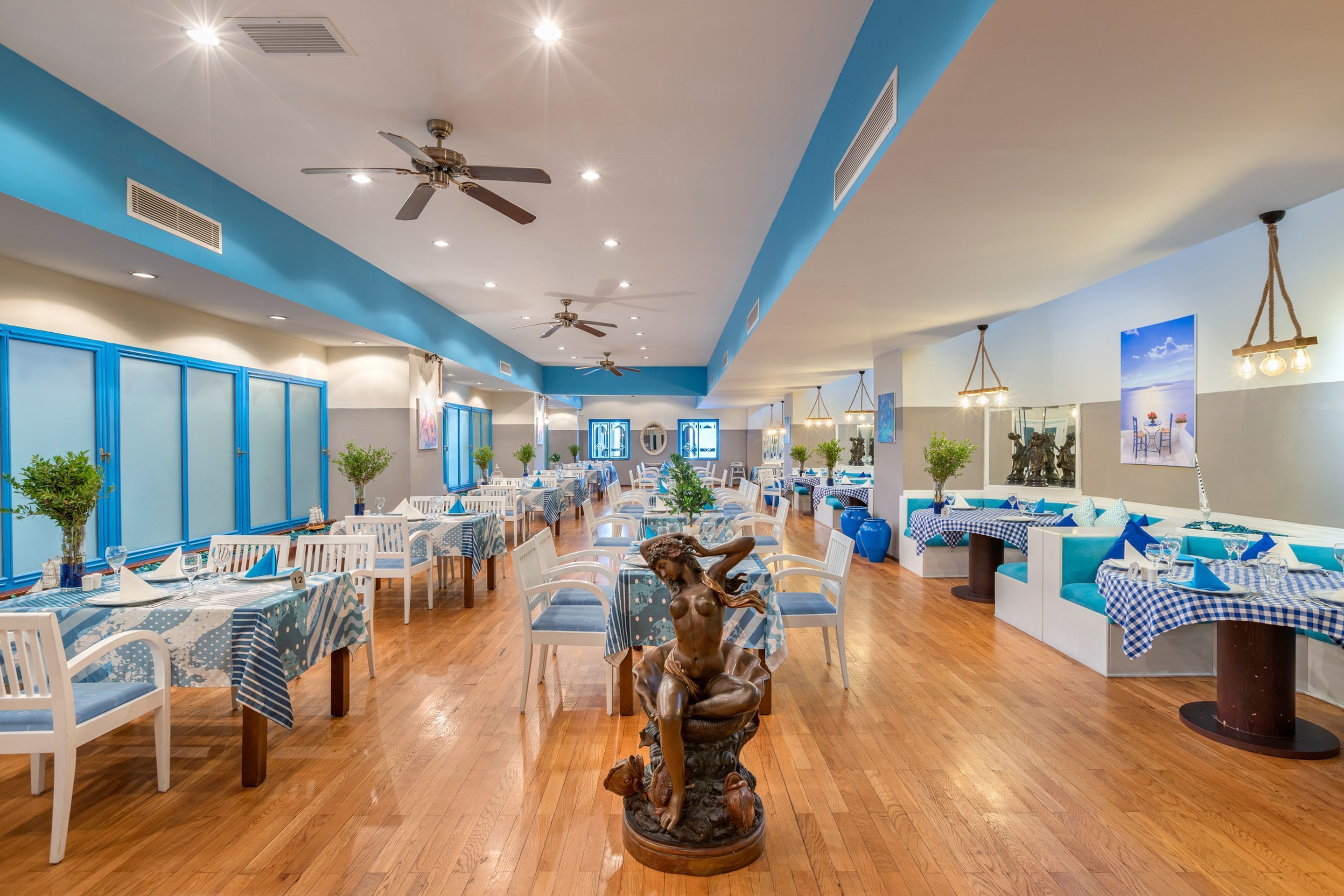 Un restaurante espacioso con techos altos, ventiladores de techo y una estatua central. Las mesas están dispuestas con manteles a cuadros azules y blancos, y hay plantas en macetas y obras de arte en las paredes.