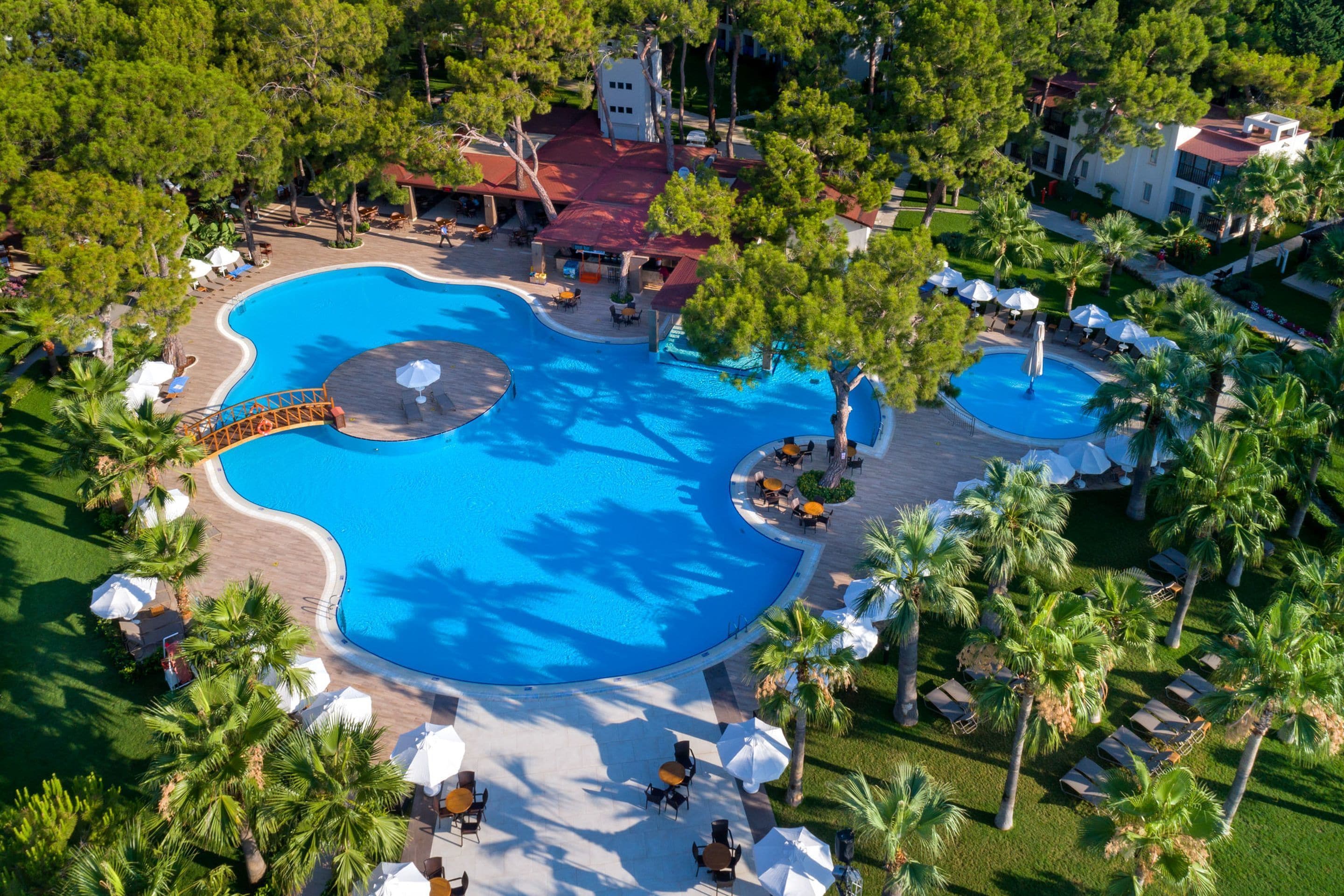 Vista aérea de un complejo turístico con una amplia piscina central, varias piscinas más pequeñas y zonas de estar al aire libre rodeadas de palmeras.