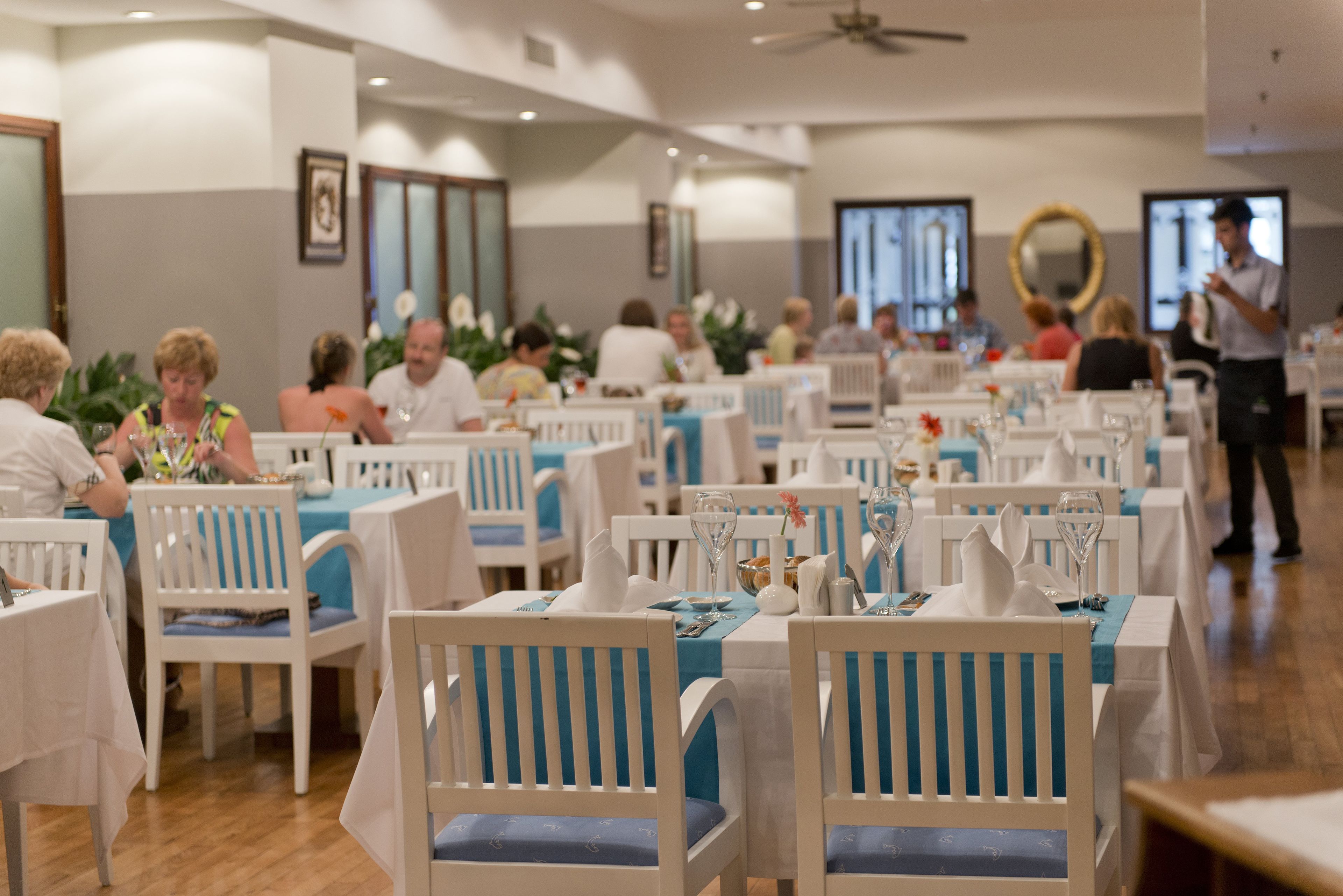Uma área de refeições com cadeiras e mesas brancas, algumas com toalhas de mesa azuis.
