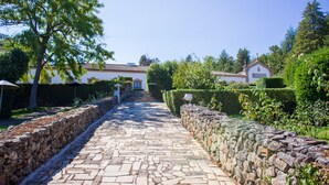 Un chemin de pierres mène à un bâtiment blanc doté d’un toit en tuiles, entouré de verdure et d’un mur de pierres.