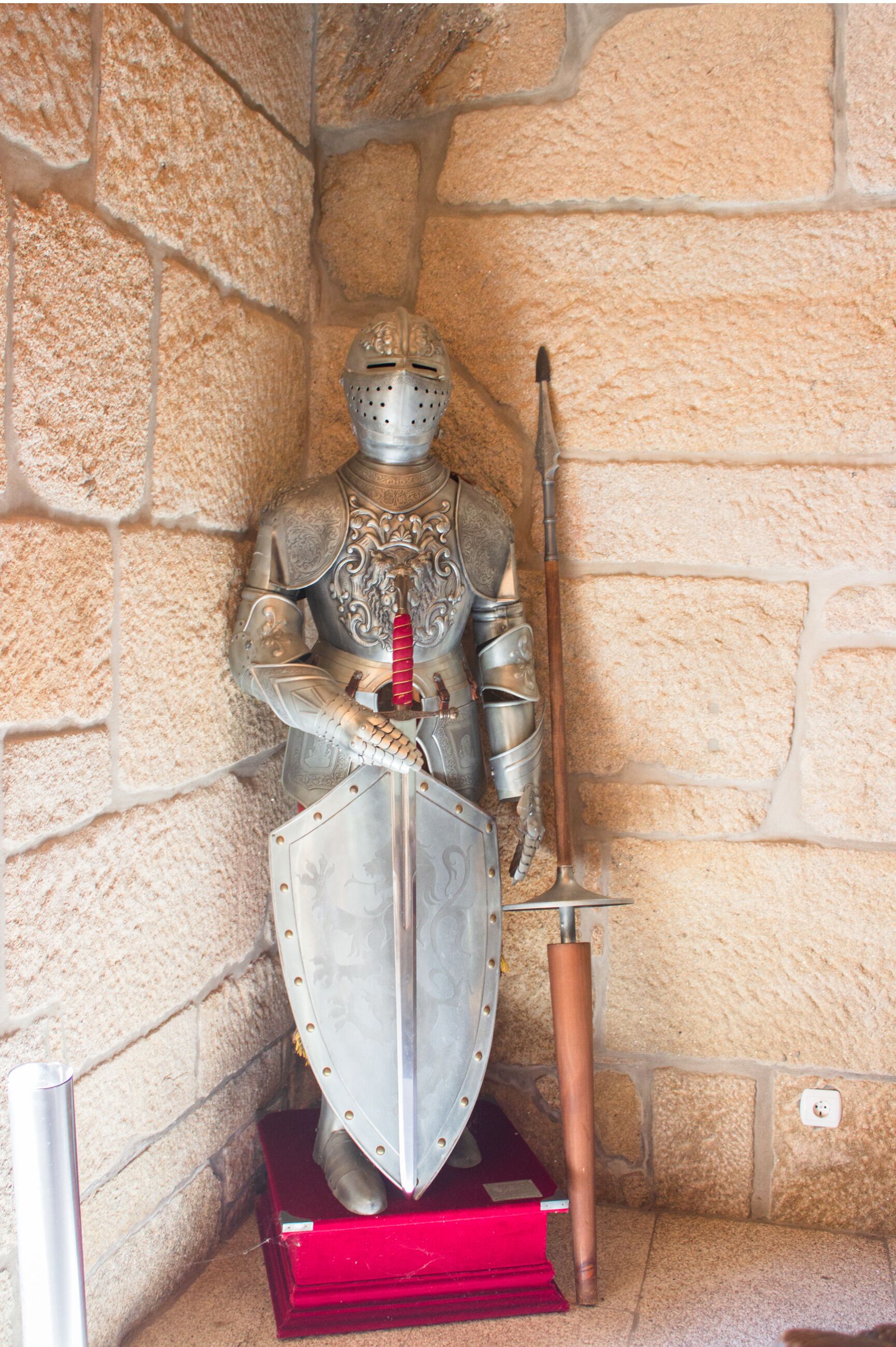 A statue of a knight in armor standing on a red pedestal against a stone wall.