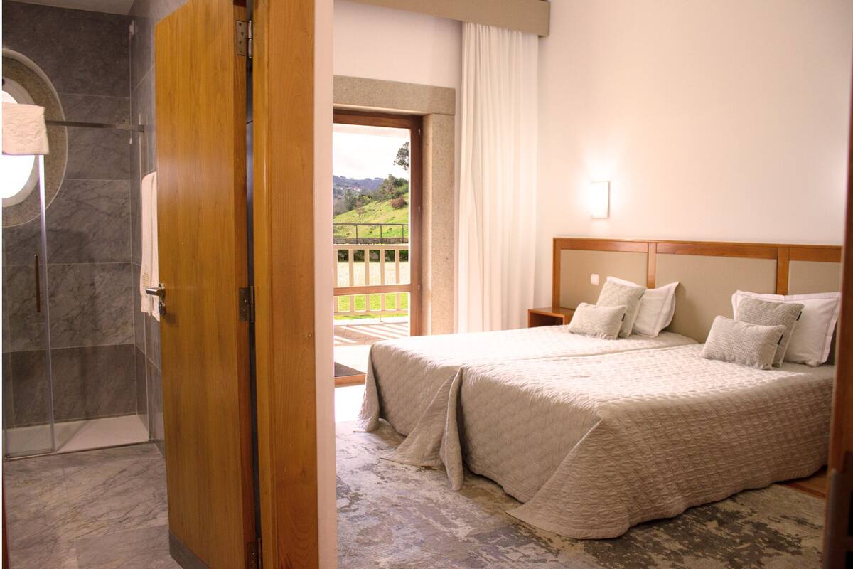 A hotel room with a large bed, a wooden headboard, a bathroom with a shower, and a balcony with a view of the countryside.