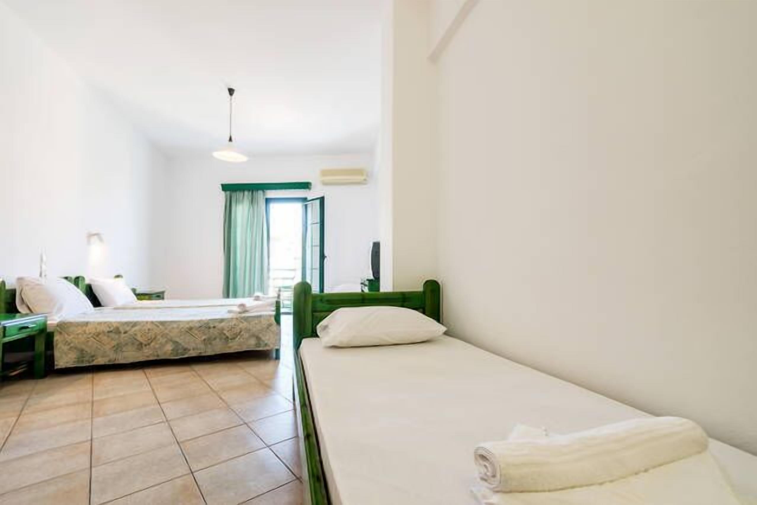A hotel room with two single beds, a green headboard, a white wall, a window with curtains, and a tiled floor.