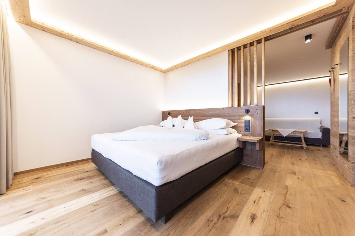 A modern bedroom with a wooden floor, a bed with white bedding, a nightstand, and a small table with a lamp.