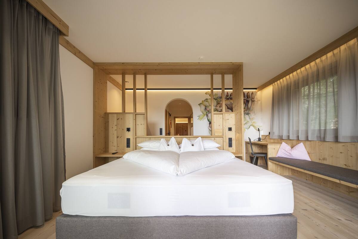 A modern bedroom with a large bed, wooden paneling, a desk, and a view of a forested area.