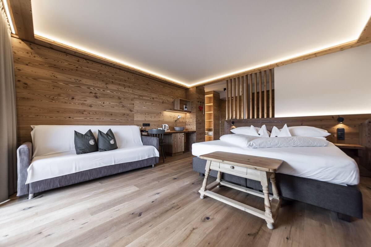 A modern hotel room with a wooden wall, a sofa, a bed, a small table, and a desk with a chair.