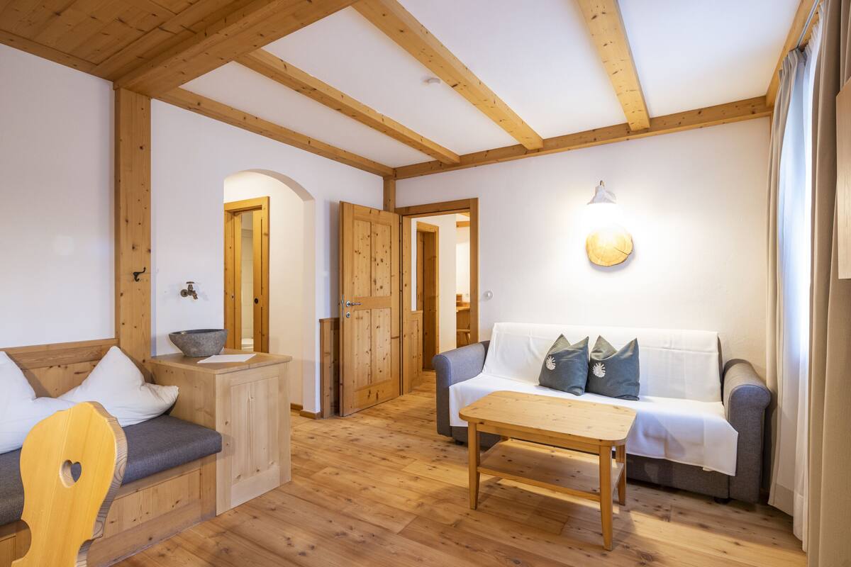 A cozy room with a wooden bench, a sofa, and a table. There is a wooden door and a light fixture on the wall.