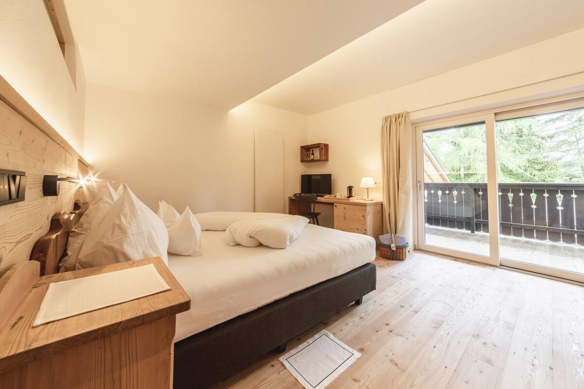 A bedroom with a bed, wooden desk, TV, and a balcony with a view of greenery.