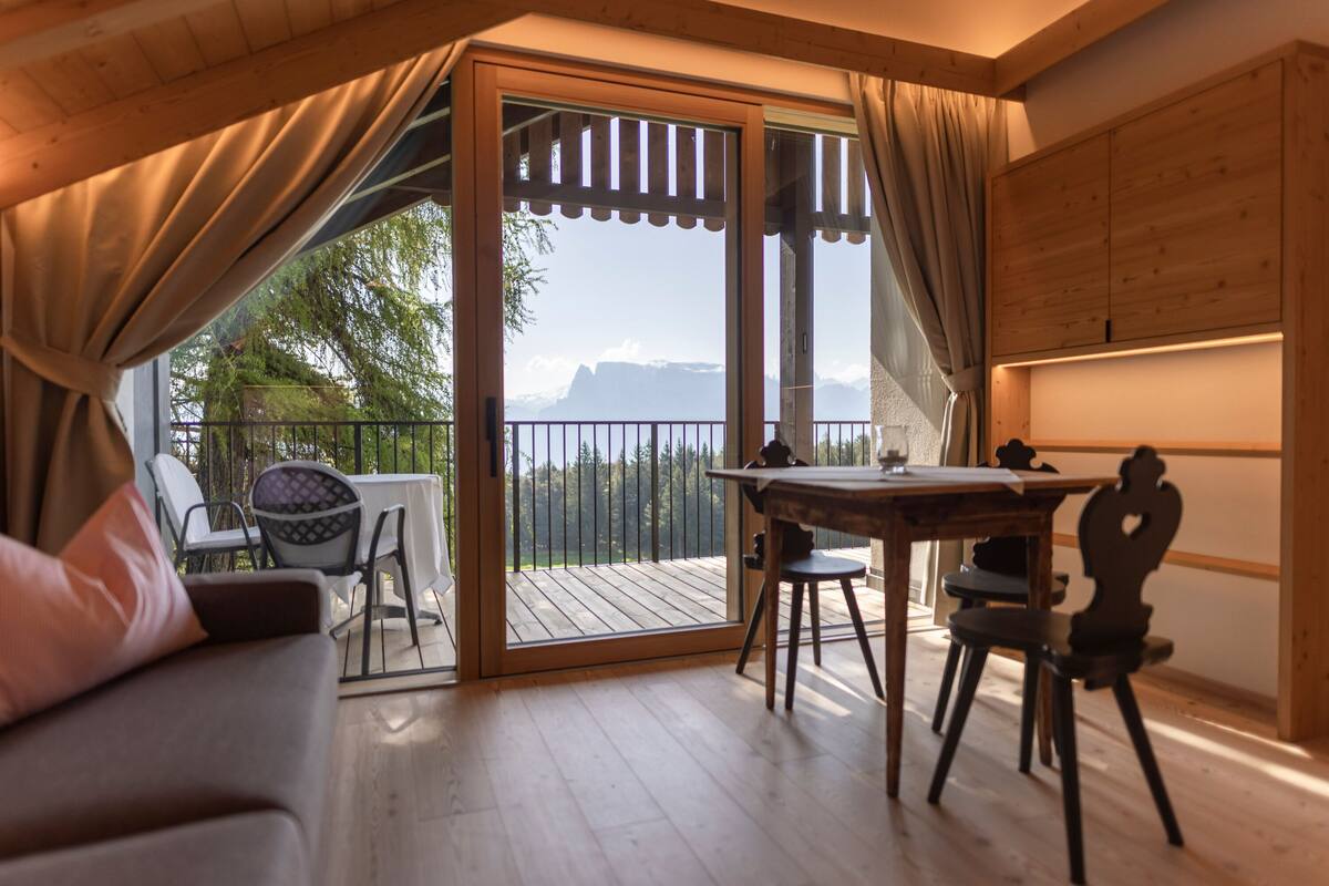 A cozy room with a wooden floor, a dining table with chairs, a sofa, and a large sliding glass door leading to a balcony with a view of mountains.