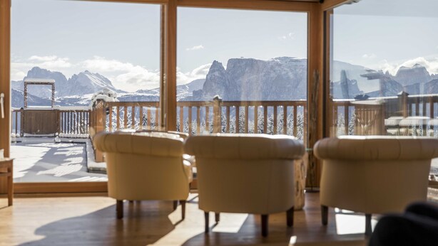 Quarto com teto de madeira e grandes janelas com vista para montanhas nevadas. Três poltronas bege e uma mesa de madeira.