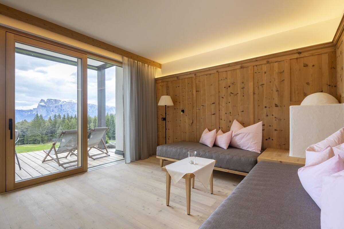 A modern room with a wooden floor, a grey sofa with pink pillows, a glass table, and a view of mountains through large glass doors.