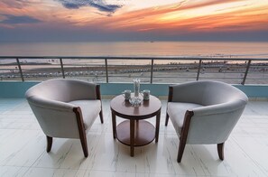A balcony with two armchairs and a small table, overlooking a beach with umbrellas and people.
