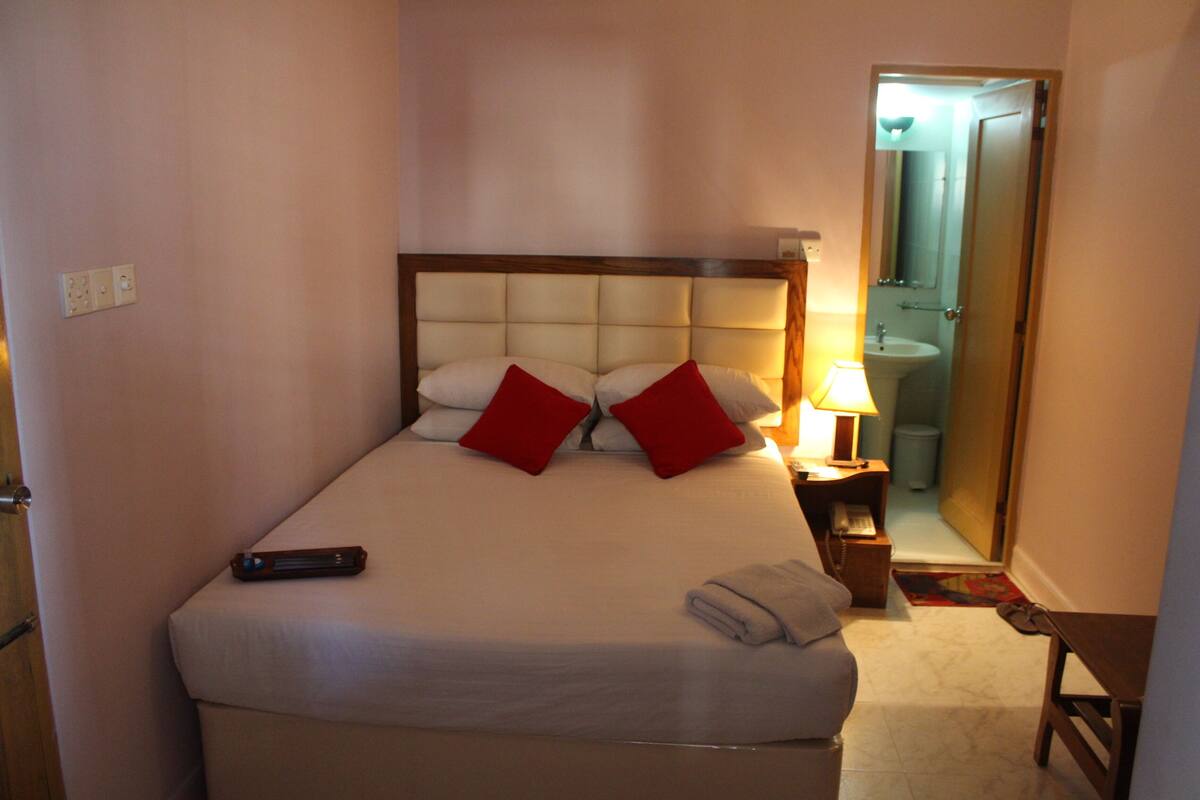 A neatly made bed with a remote control, red pillows, and a folded towel on the side.