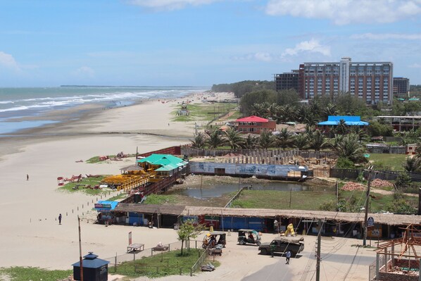 A beachside view with a sandy beach, a small water body, and a row of buildings including a hotel with a red roof.