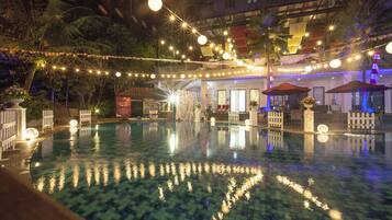 A swimming pool with a tiled pattern, surrounded by palm trees and a building with a balcony.