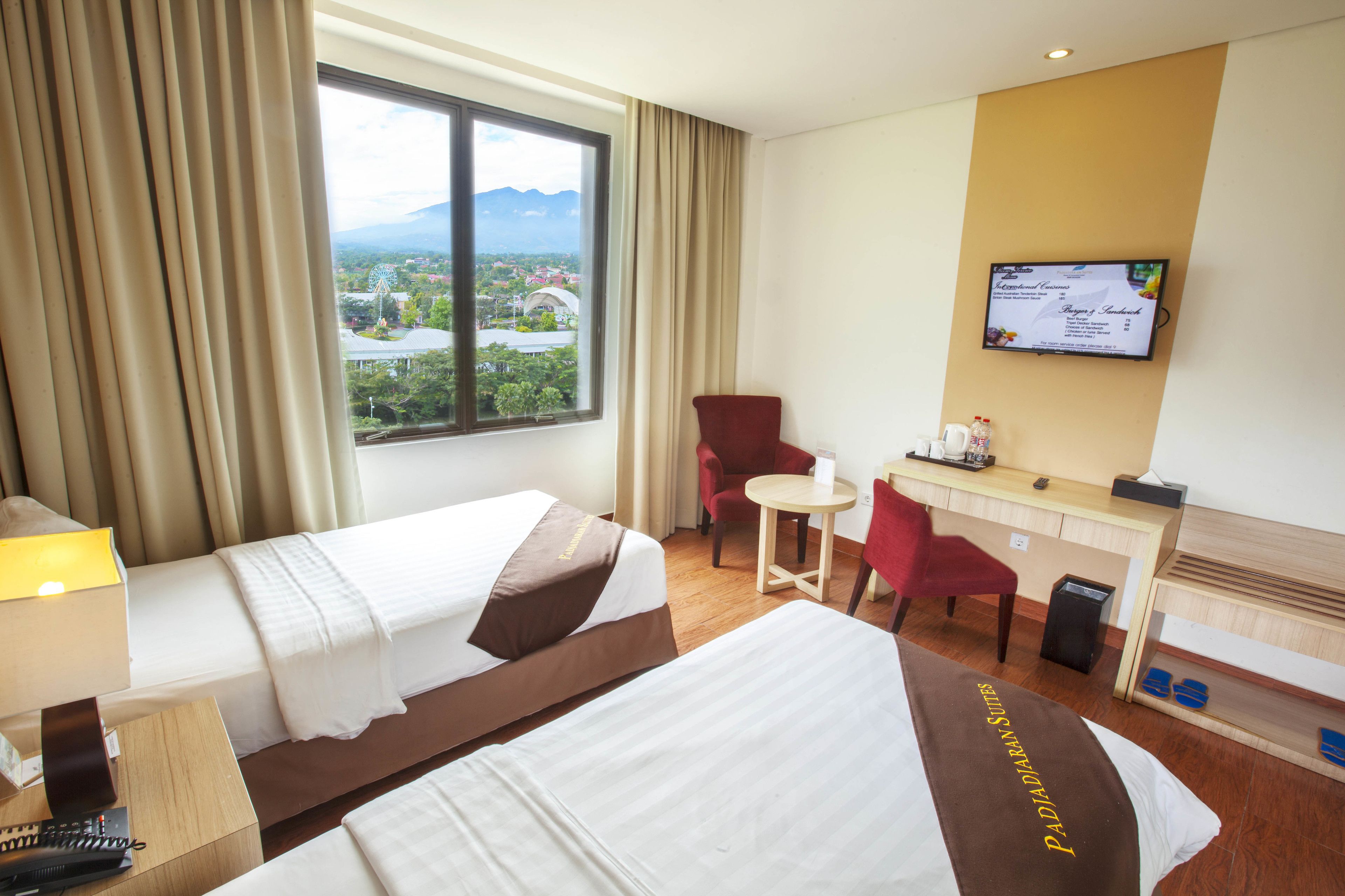 Habitación de hotel con dos camas, un escritorio, una silla, un televisor y una ventana con vistas a una ciudad y montañas.