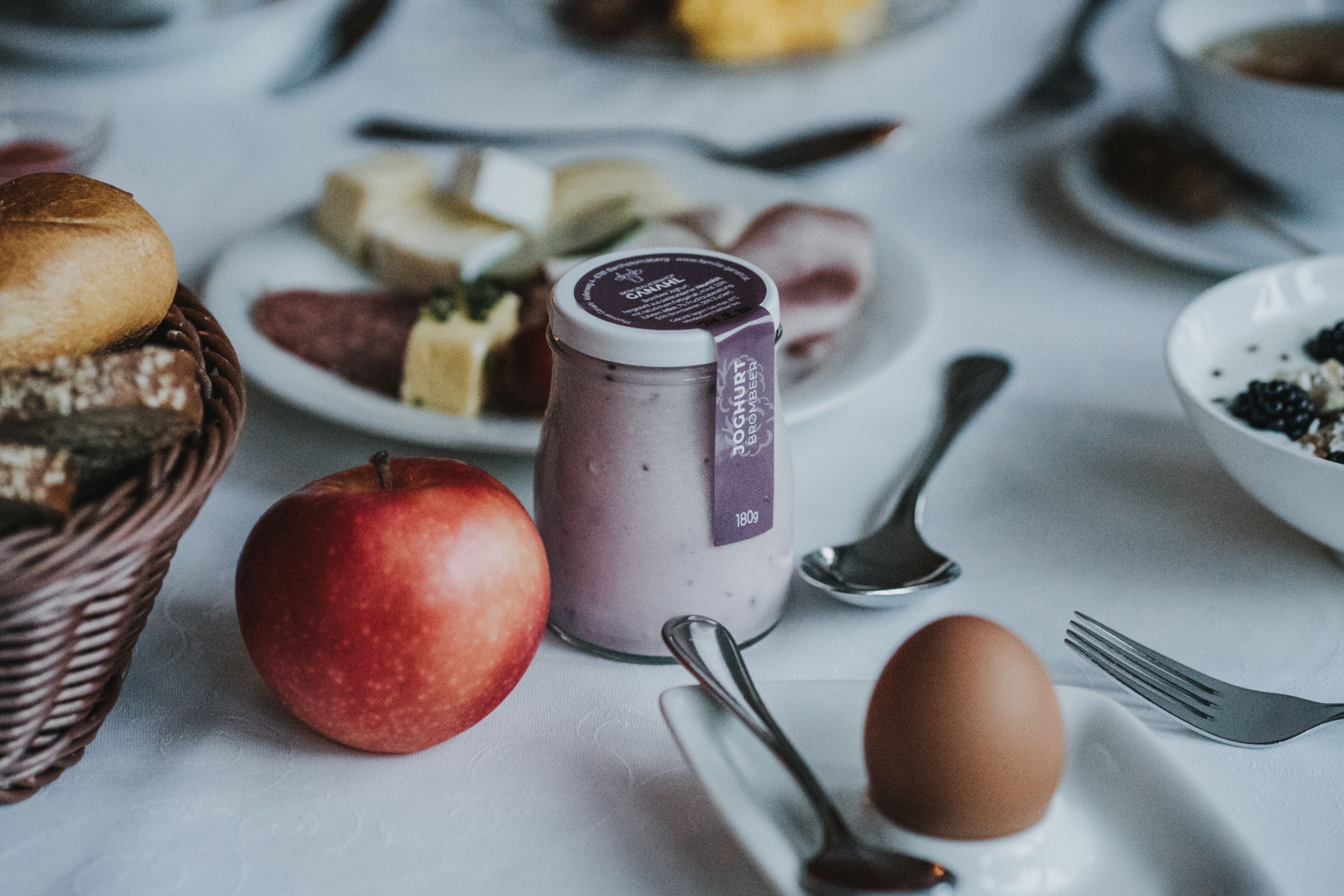 Une table sur laquelle se trouvent divers aliments, notamment une pomme, un pot de yaourt, ainsi que différentes sortes de pain et de fromage.