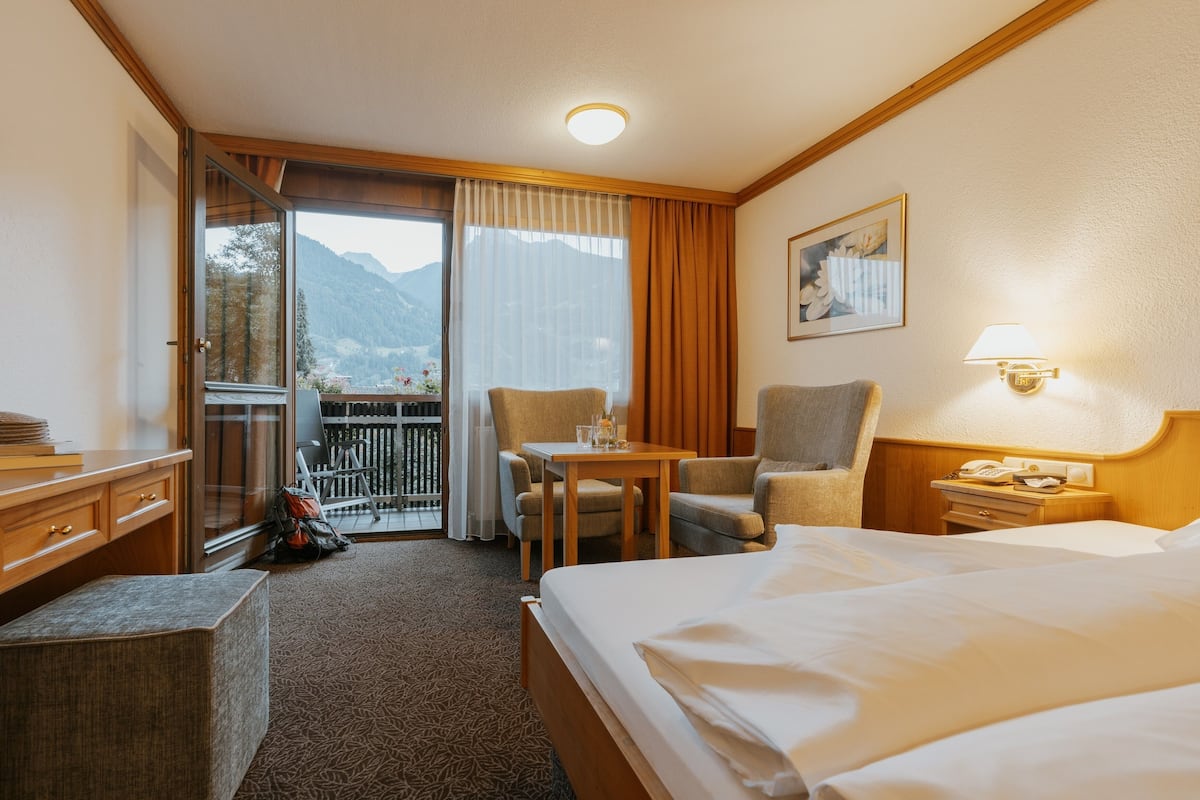 A hotel room with a bed, a desk, a chair, a small table, a nightstand, a lamp, a window with curtains, a door, and a view of mountains.