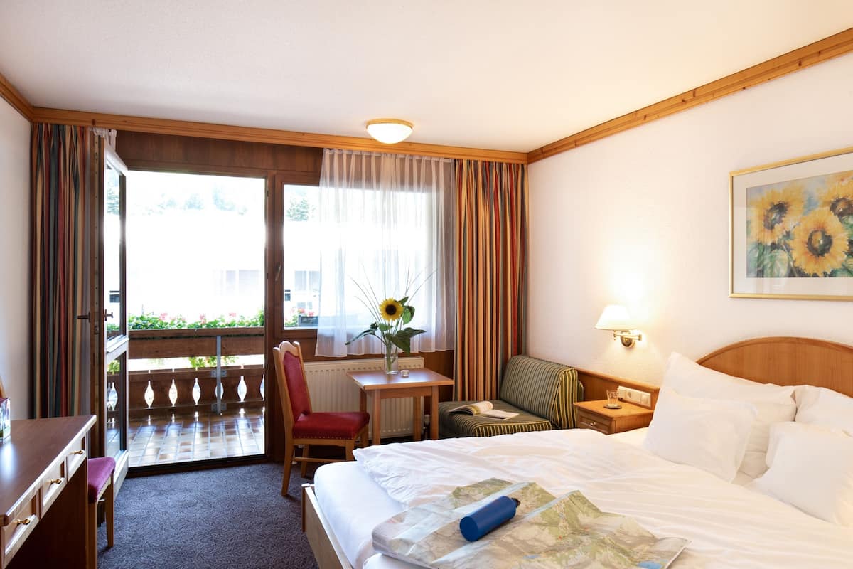 A hotel room with a bed, a desk, a chair, a table with a vase of flowers, and a window with curtains.