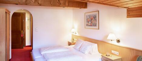 Habitación de hotel con techo de madera, una cama con ropa blanca, dos lámparas de noche, una mesita de noche con un vaso y un cuadro en la pared.