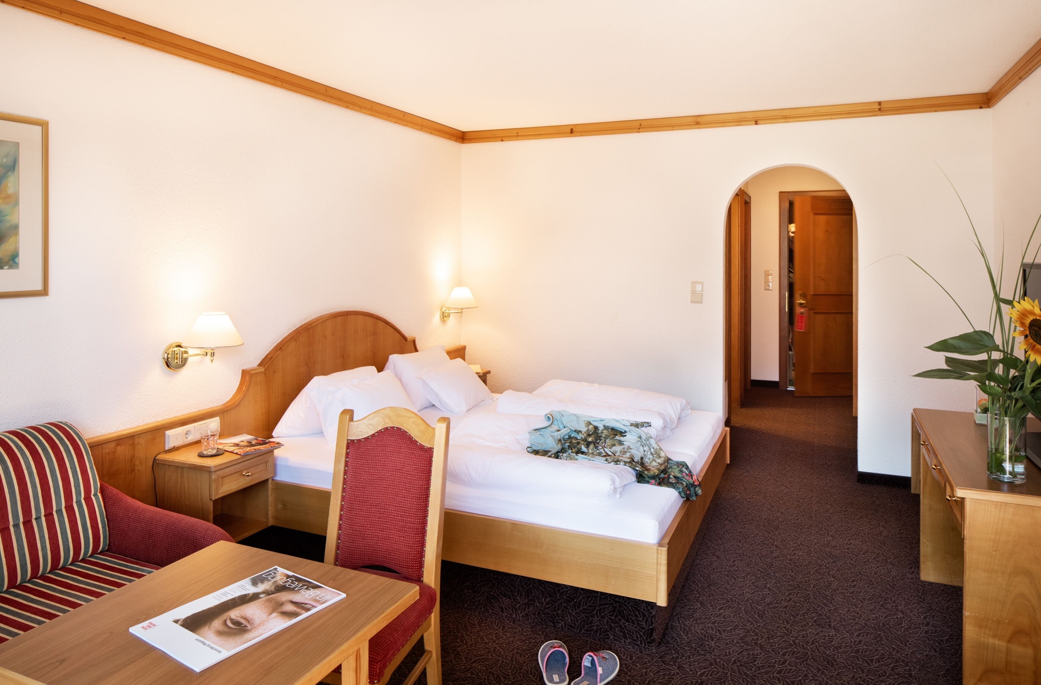 Una habitación de hotel con una cama, una mesita de noche de madera, una silla roja, una mesa de madera y un escritorio pequeño con una lámpara.
