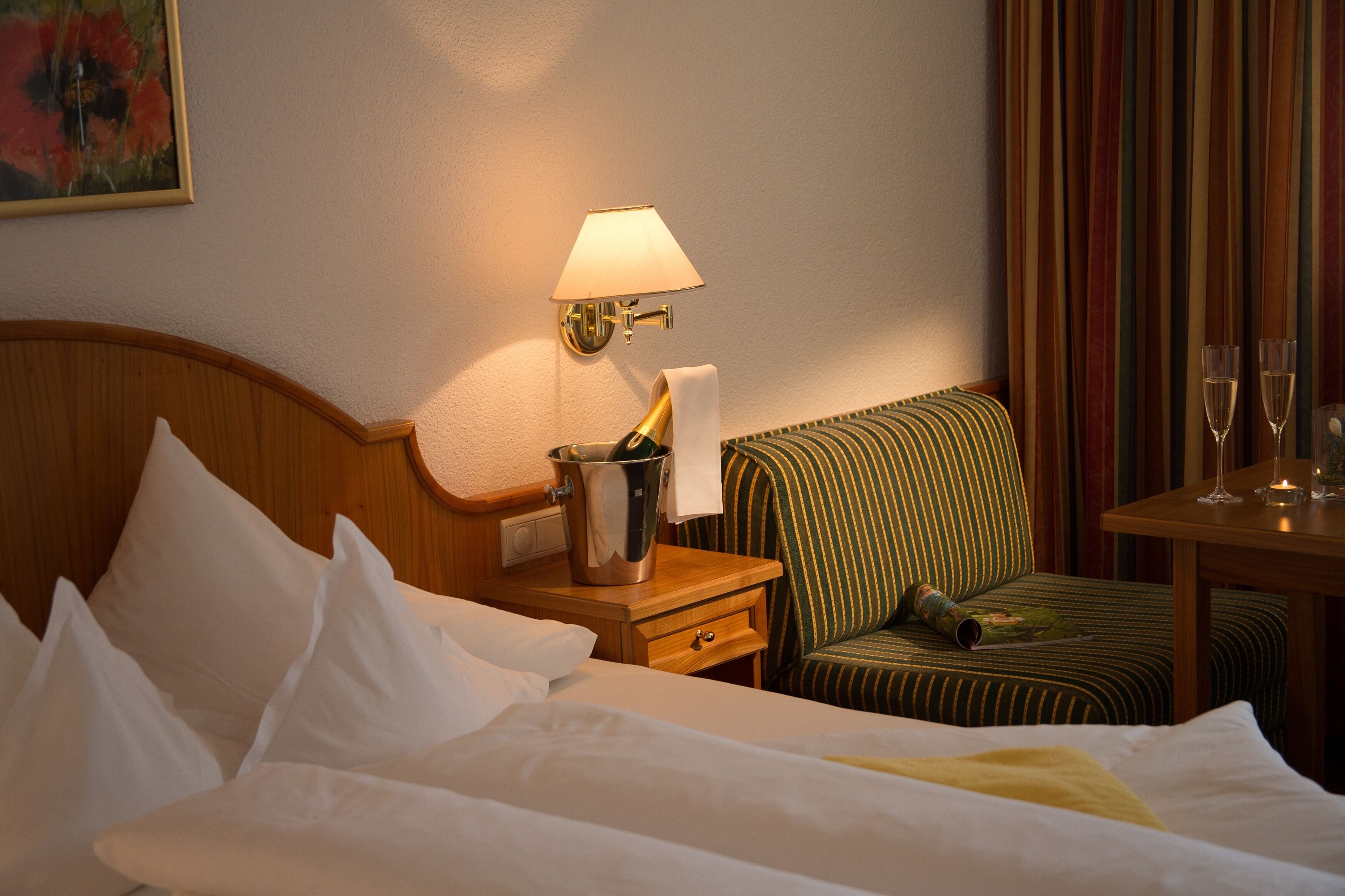 A hotel room with a bed, a nightstand with a lamp, a chair, and a table with glasses and a bottle.