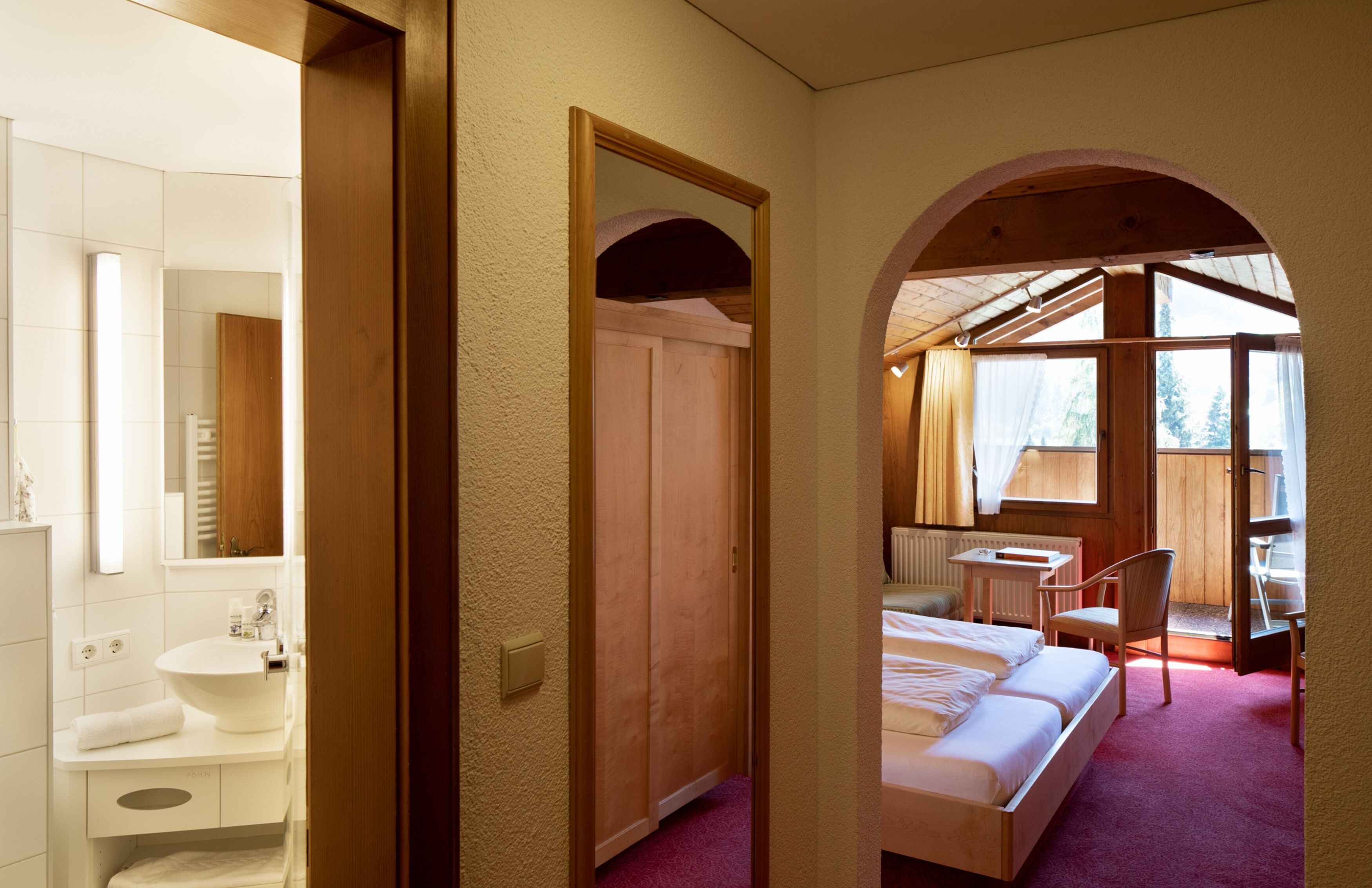 Quarto com cama, escrivaninha e cadeira. Inclui banheiro com pia e espelho. O quarto possui paredes de madeira e porta em arco.