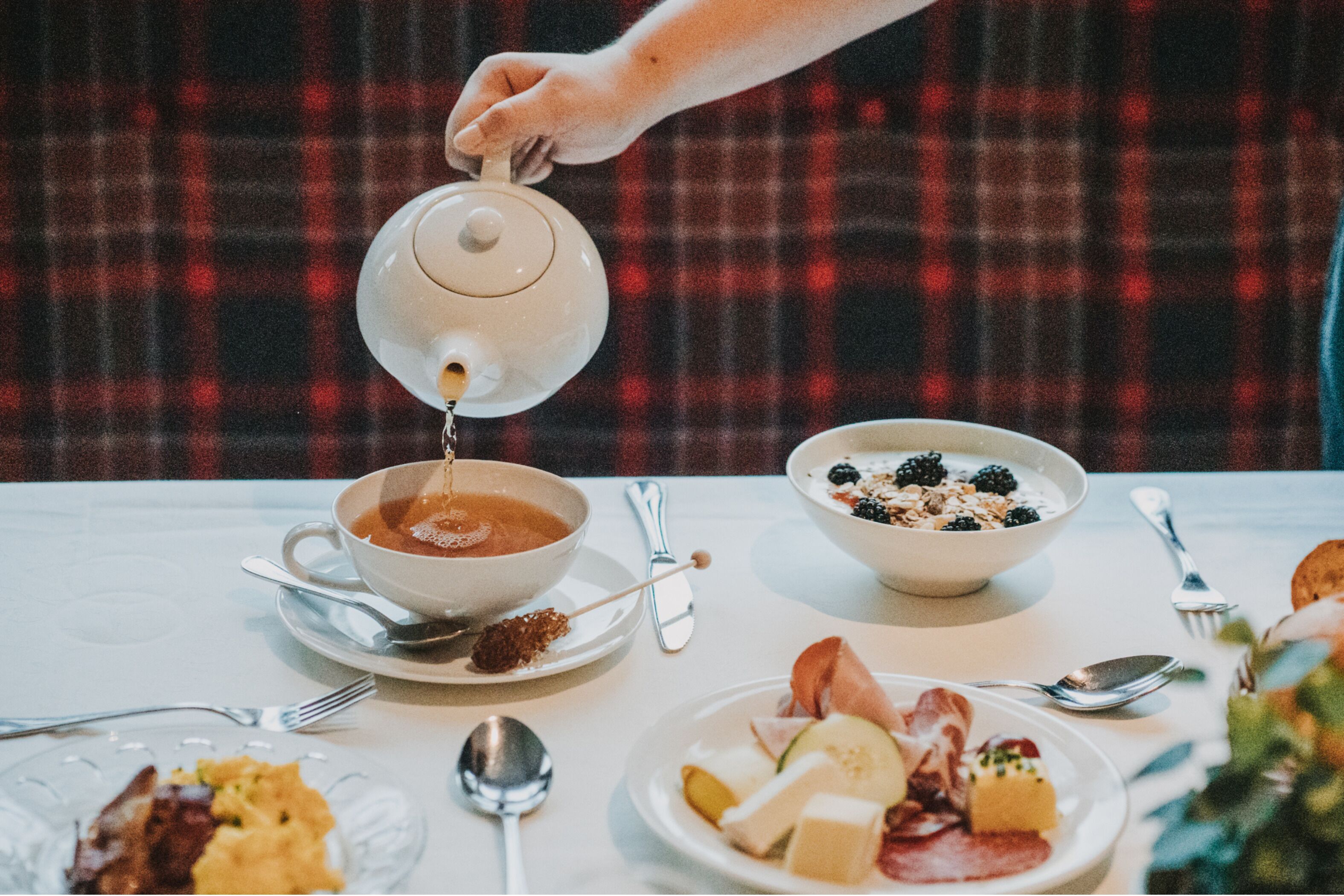 A breakfast setting with a teapot pouring into a cup, a bowl of cereal, and plates with various foods.