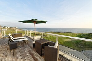 A wooden deck with wicker furniture and a green umbrella overlooking a coastal area. - Hotel Le Lichen (Batz-sur-Mer)