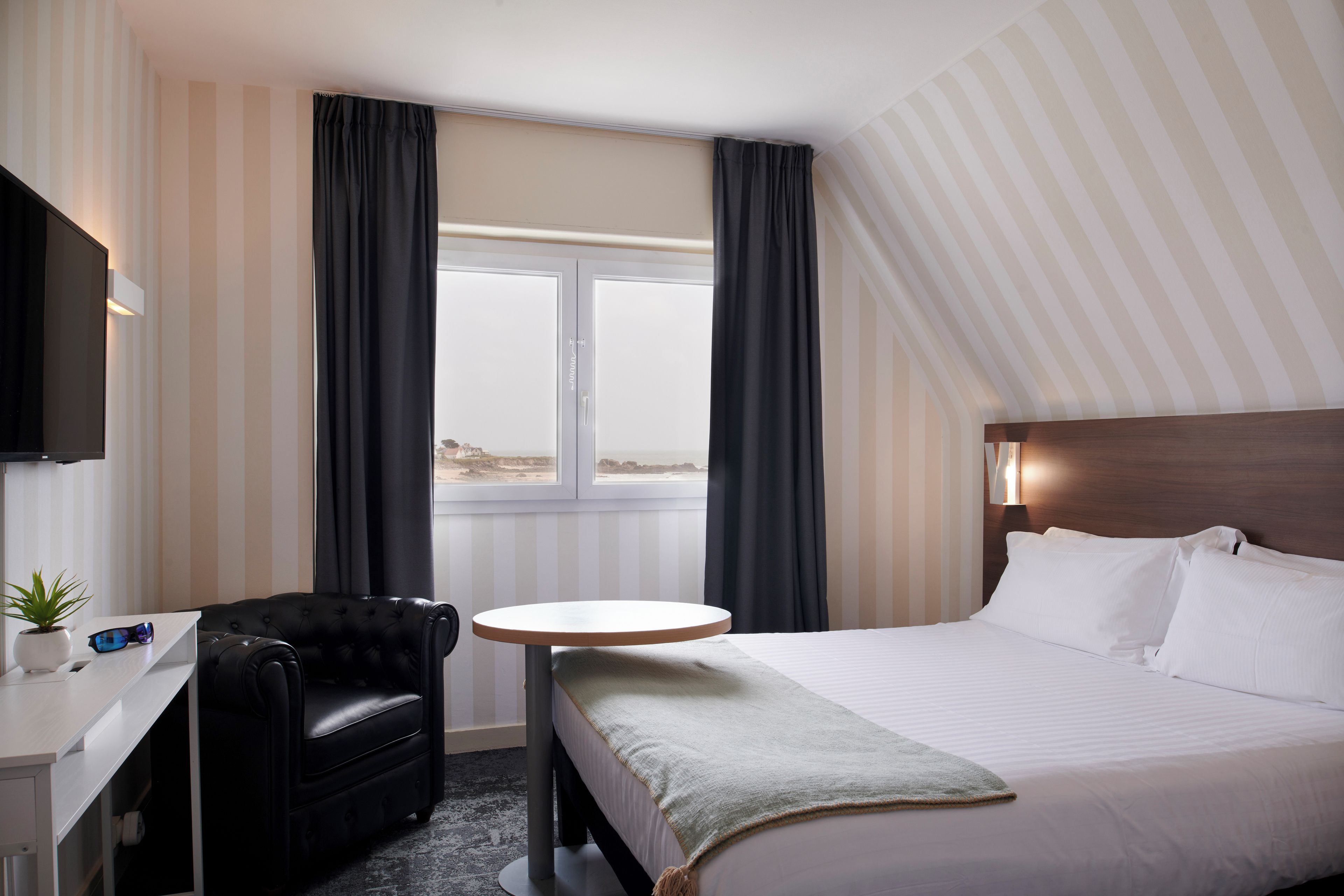 A hotel room with a bed, a desk with a chair, a small round table, and a TV mounted on the wall.