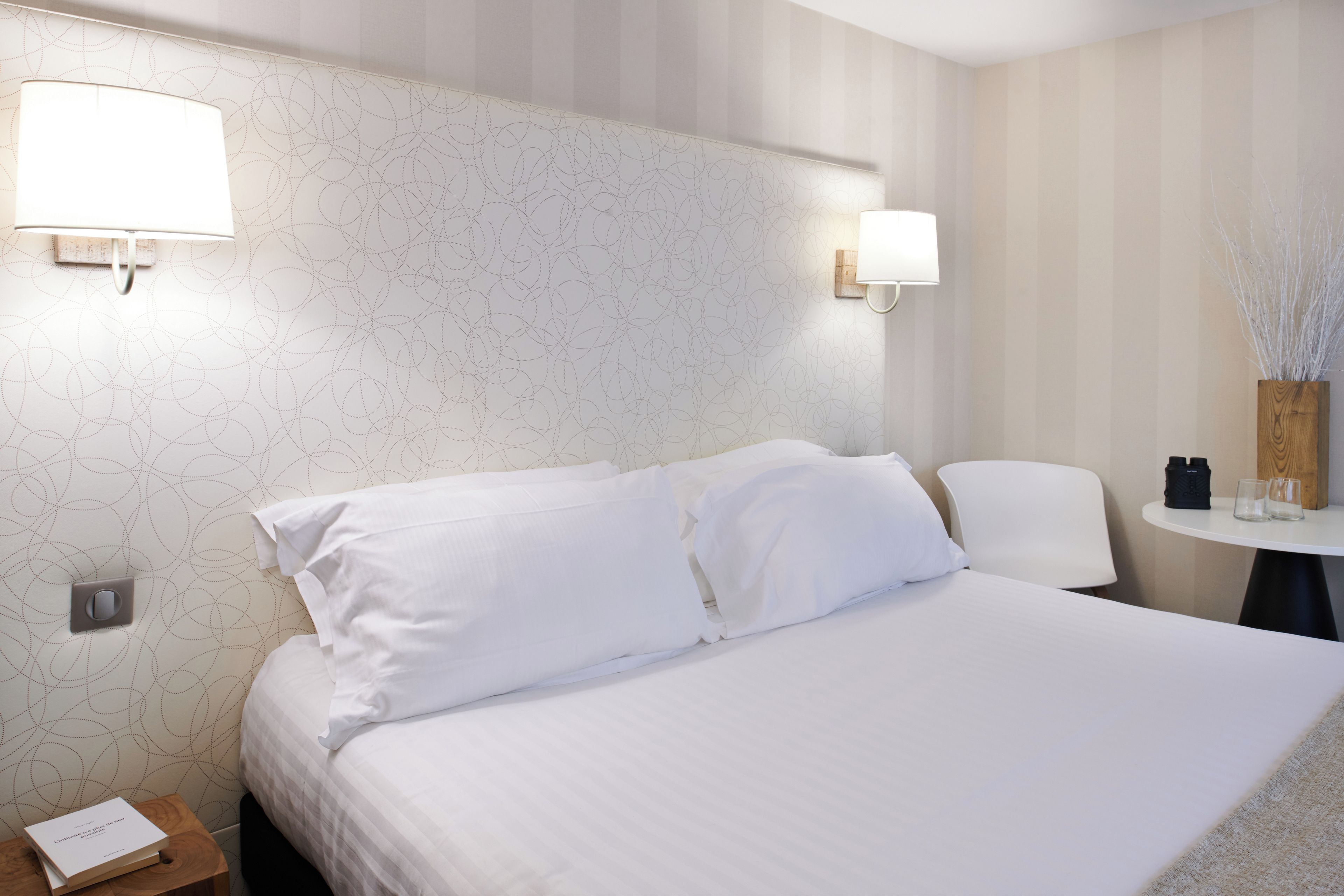 A hotel room with a bed, two bedside lamps, a small table with a vase, and a chair.