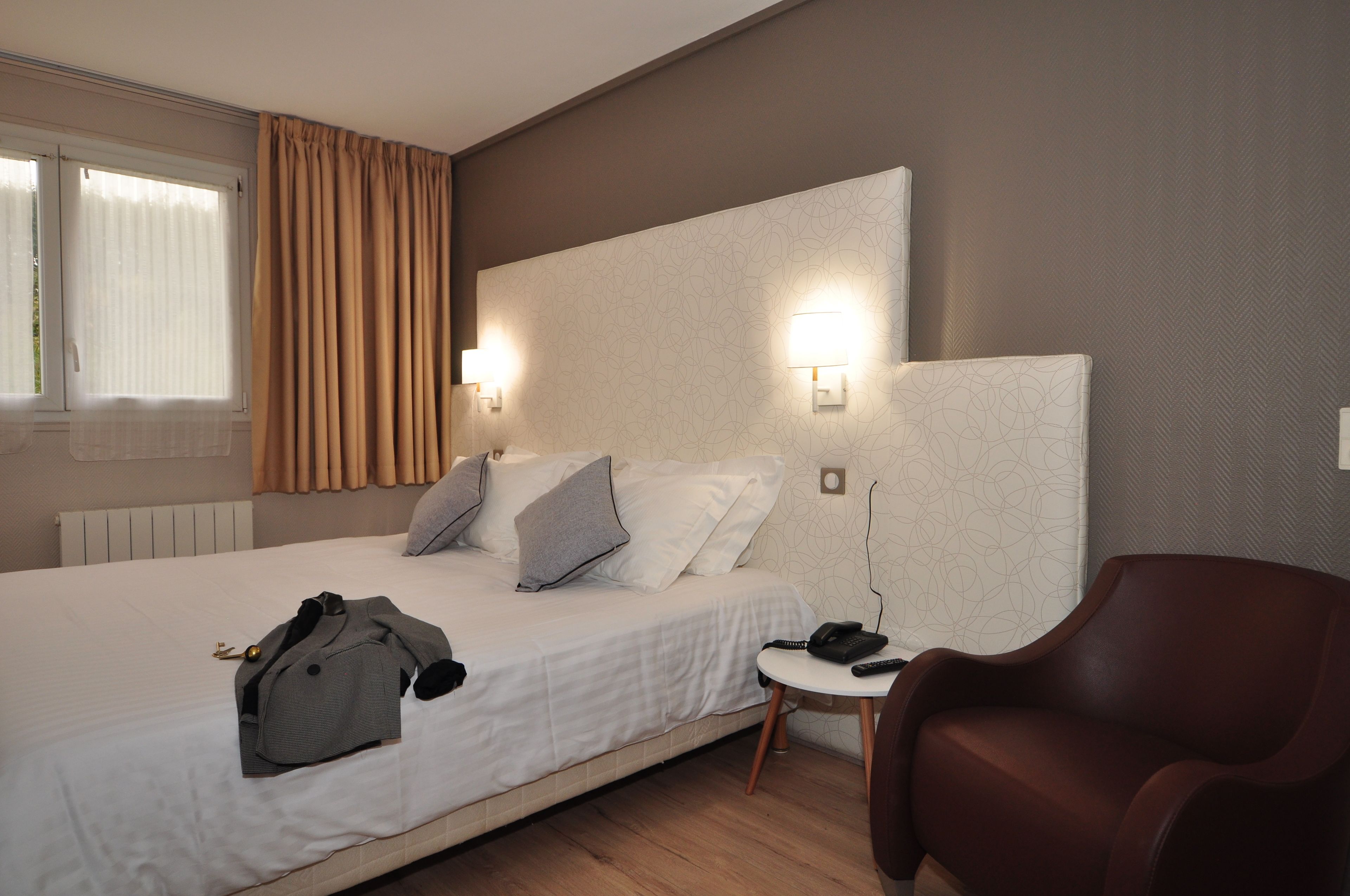 A hotel room with a bed, a chair, a bedside table, and a window with curtains.