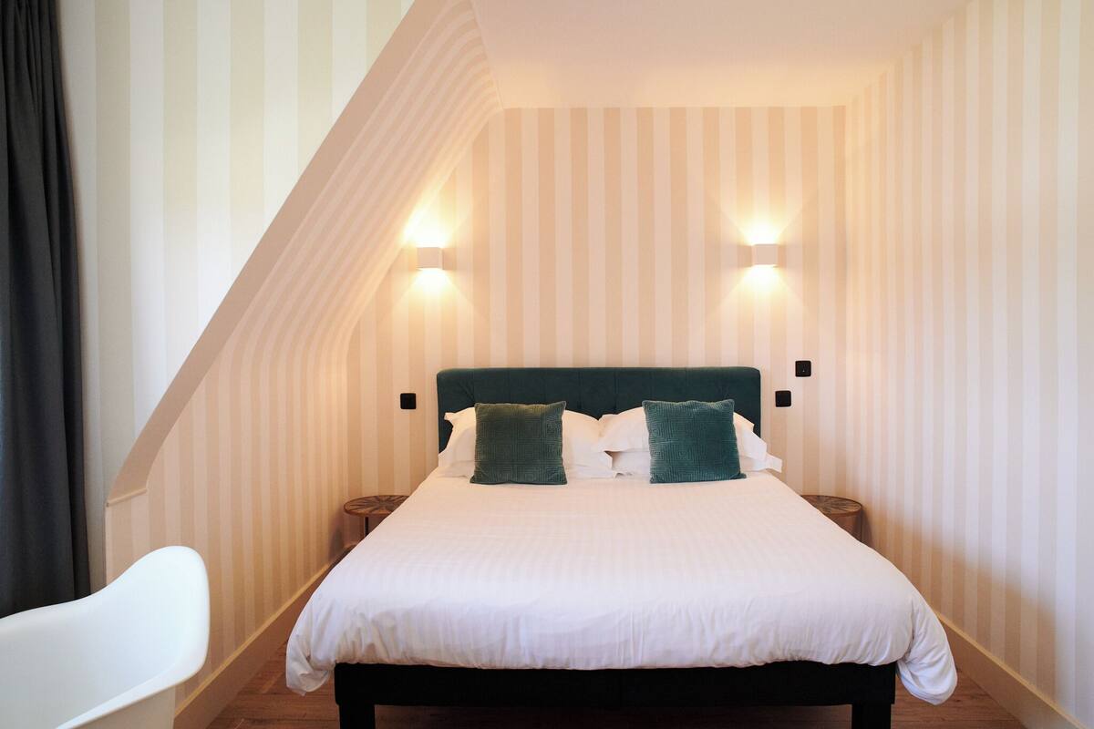 A neatly made bed with white bedding and green pillows in a room with striped wallpaper and two wall-mounted lights.
