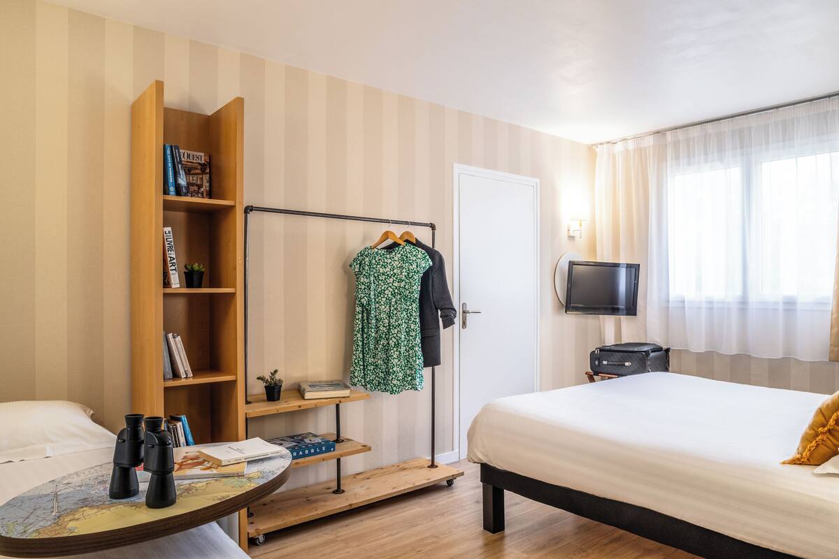 A hotel room with a bed, a wooden bookshelf, a coat rack, a television, and a small table with a map and binoculars.