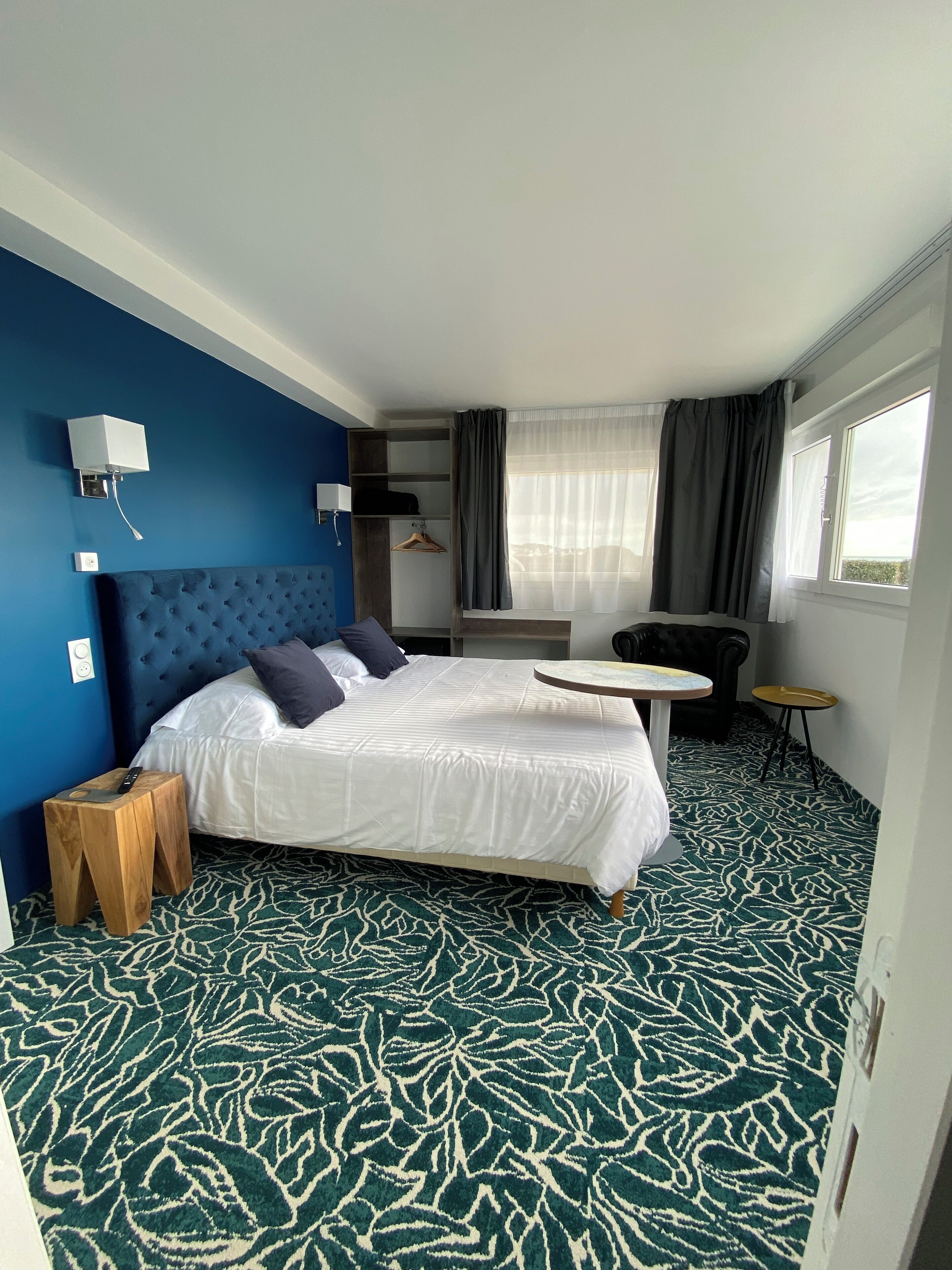 Una habitación de hotel con una cama grande, una mesita, una silla, un armario y un ventanal con cortinas.