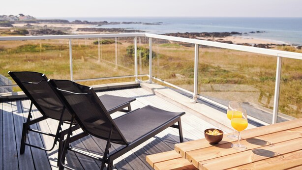 A balcony with two black lounge chairs, a wooden table with two glasses of orange juice, and a bowl of chips. - Hotel Le Lichen (Batz-sur-Mer)