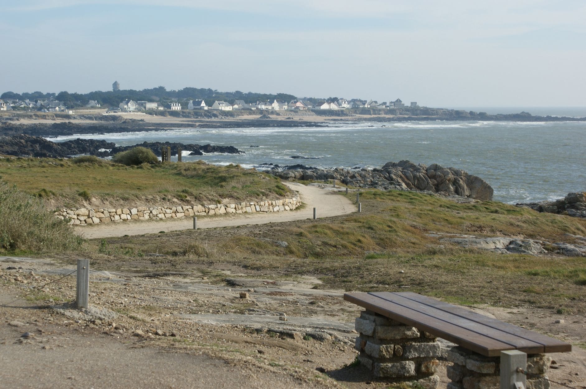 A coastal path with a stone bench, leading to a rocky shoreline with waves.