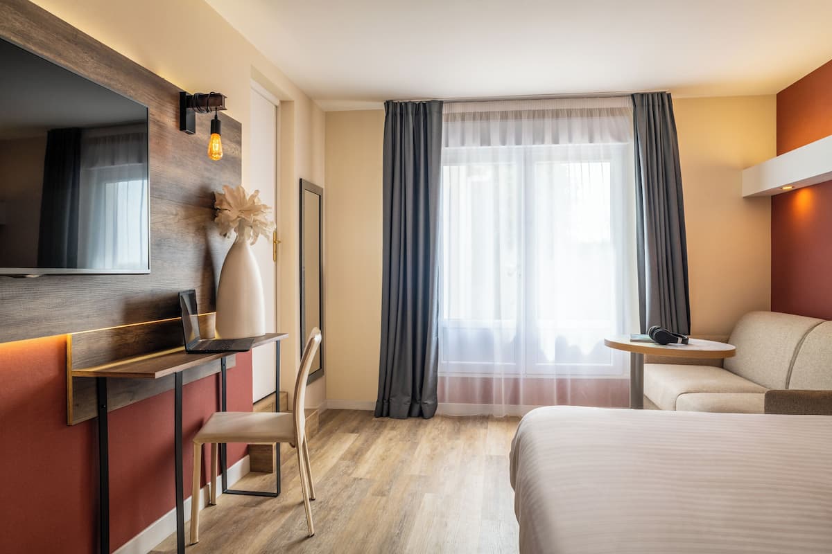 A hotel room with a bed, a desk with a laptop, a chair, a TV, and a window with curtains.