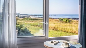 A room with a large window overlooking a coastal landscape, a small table with a cup and a book, and a map. - Hotel Le Lichen (Batz-sur-Mer)