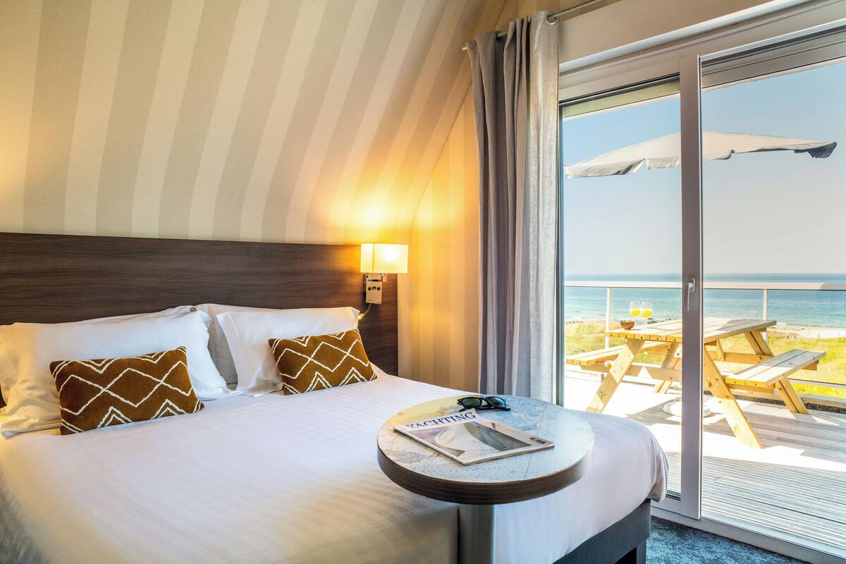 A hotel room with a bed, a bedside table with a magazine, and a view of the beach through a sliding glass door.