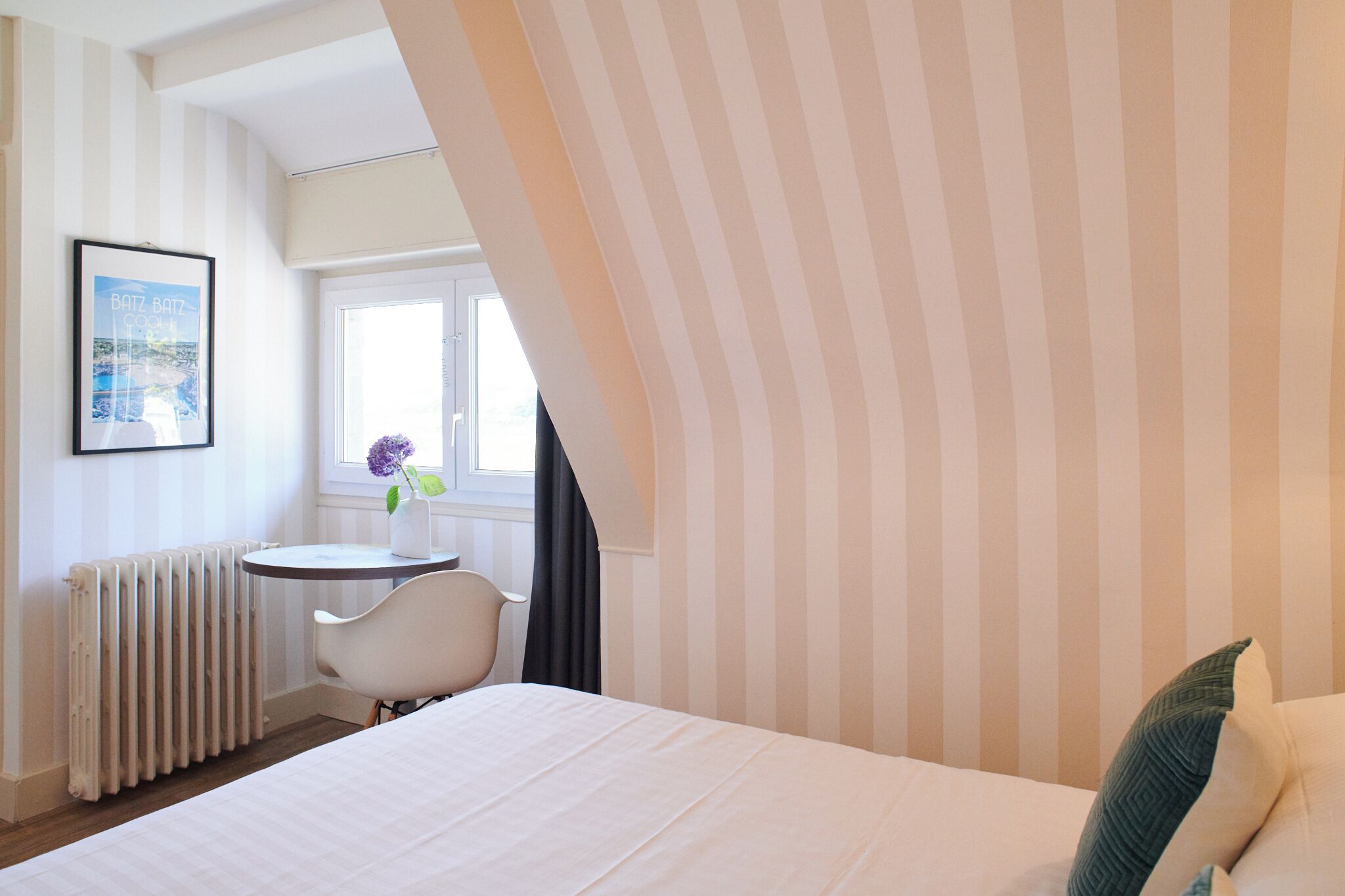 Habitación de hotel con cama, una mesita pequeña con una silla, un radiador, una ventana con cortinas y un cuadro en la pared.