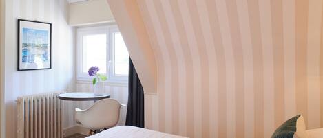 Habitación de hotel con cama, una mesita pequeña con una silla, un radiador, una ventana con cortinas y un cuadro en la pared.