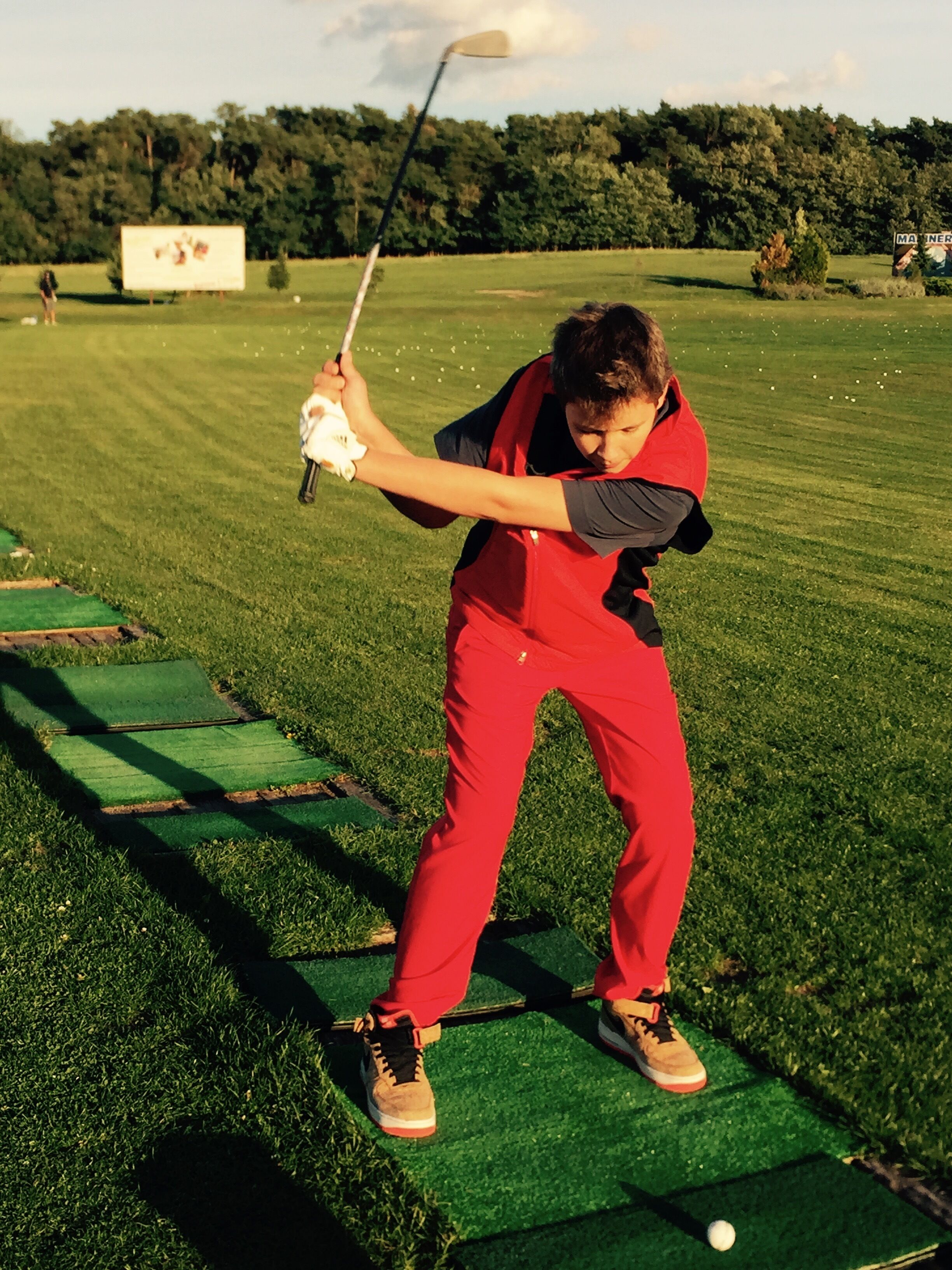 A person in red attire playing golf on a green field with trees in the background.