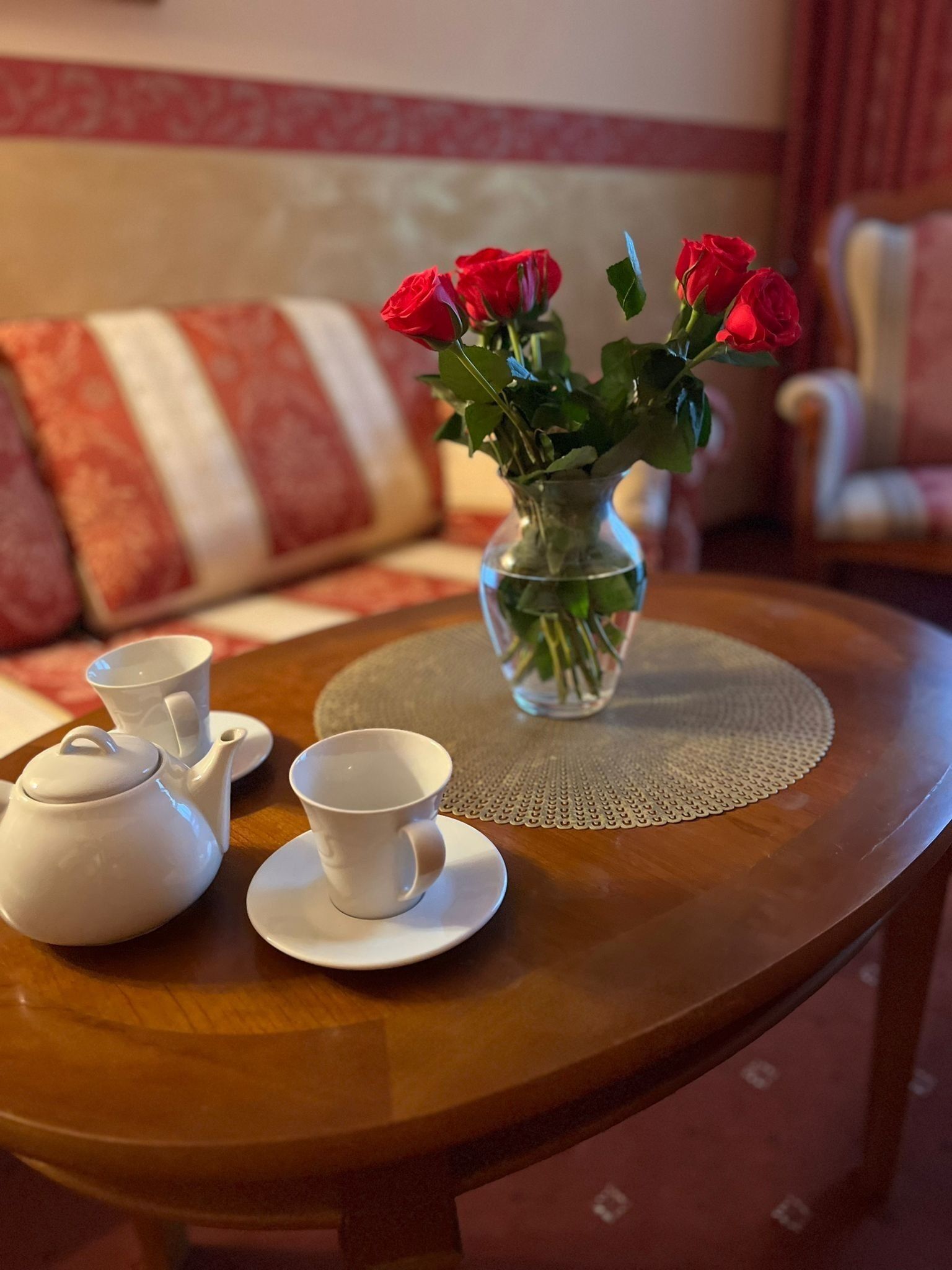 A round wooden table with a clear glass vase containing red roses, a white teapot, and two white cups on a doily.