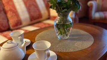 A round wooden table with a clear glass vase containing red roses, a white teapot, and two white cups on a doily.
