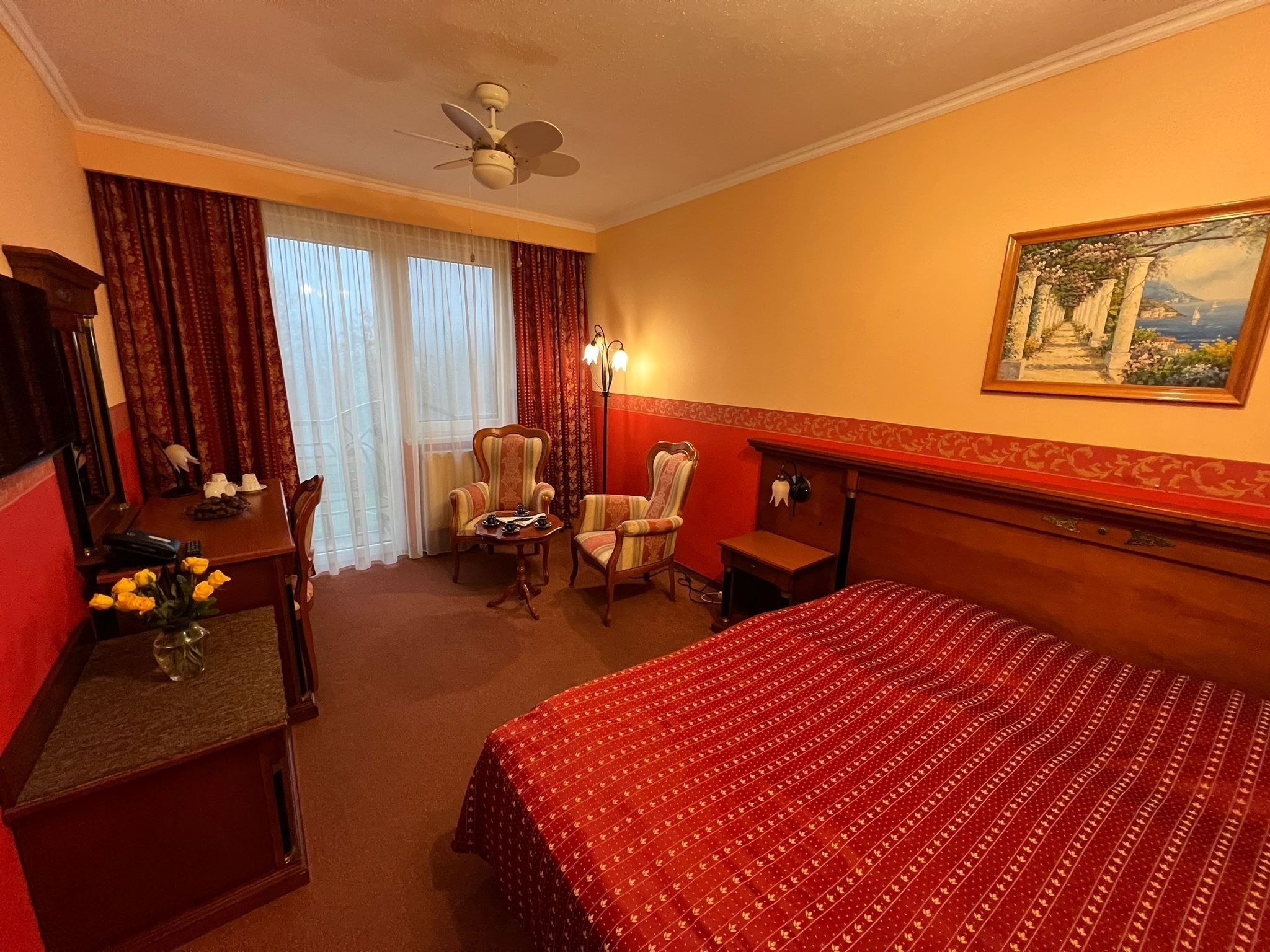 Una habitació d'hotel amb un llit, dues butaques, una taula de nit amb un telèfon i un ram de flors, un ventilador de sostre i un quadre a la paret.