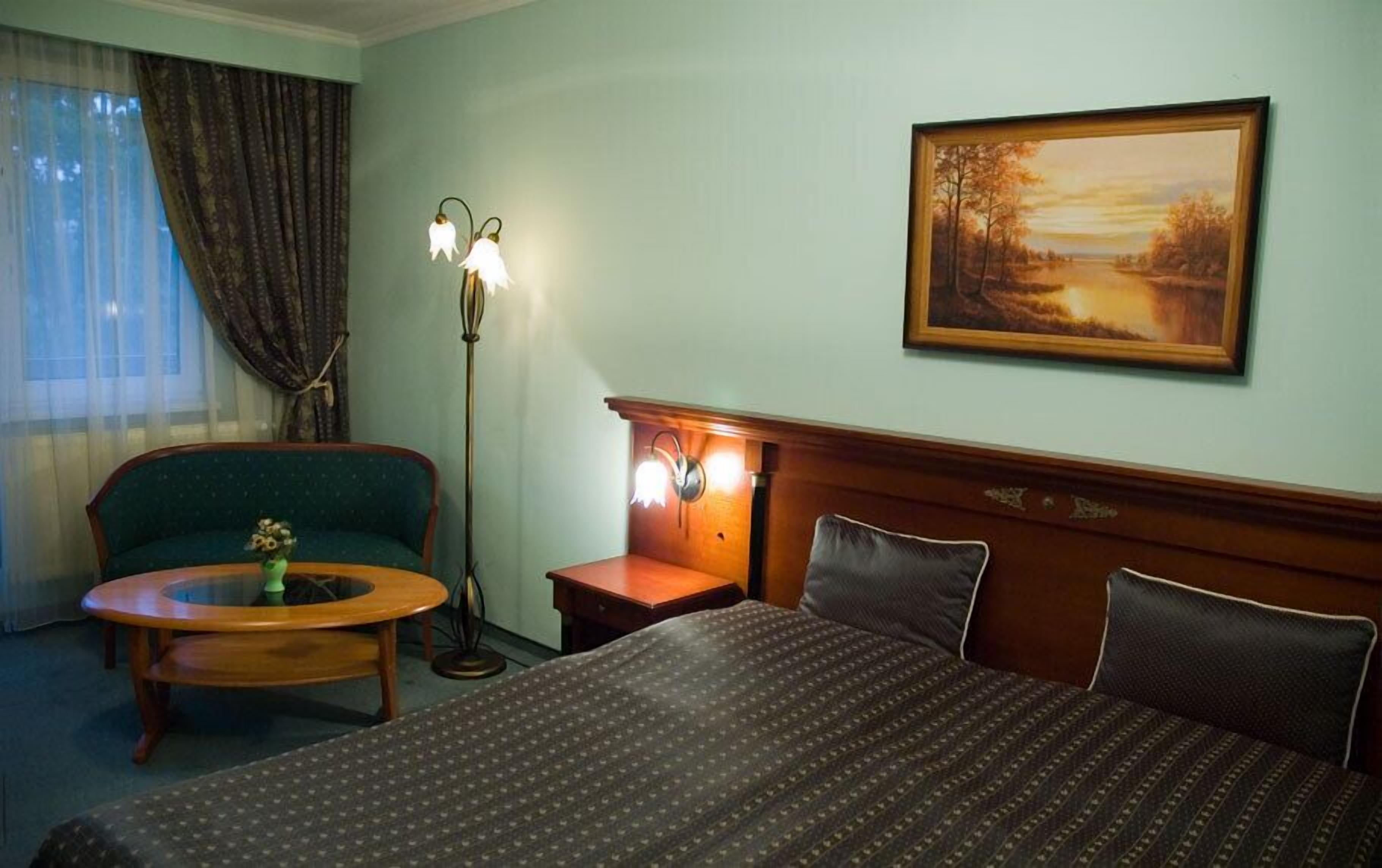 A hotel room with a bed, a chair, a small table with a vase, a floor lamp, and a painting on the wall.