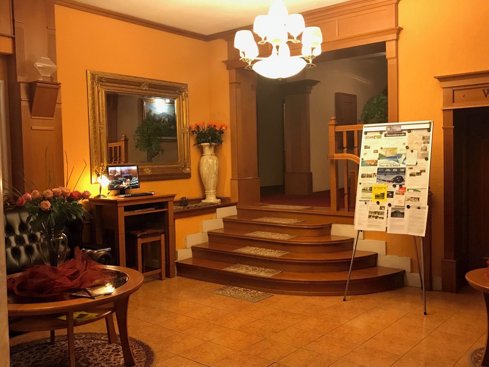 A lobby with a grand staircase, a reception desk, a large mirror, and a bulletin board with various notices.