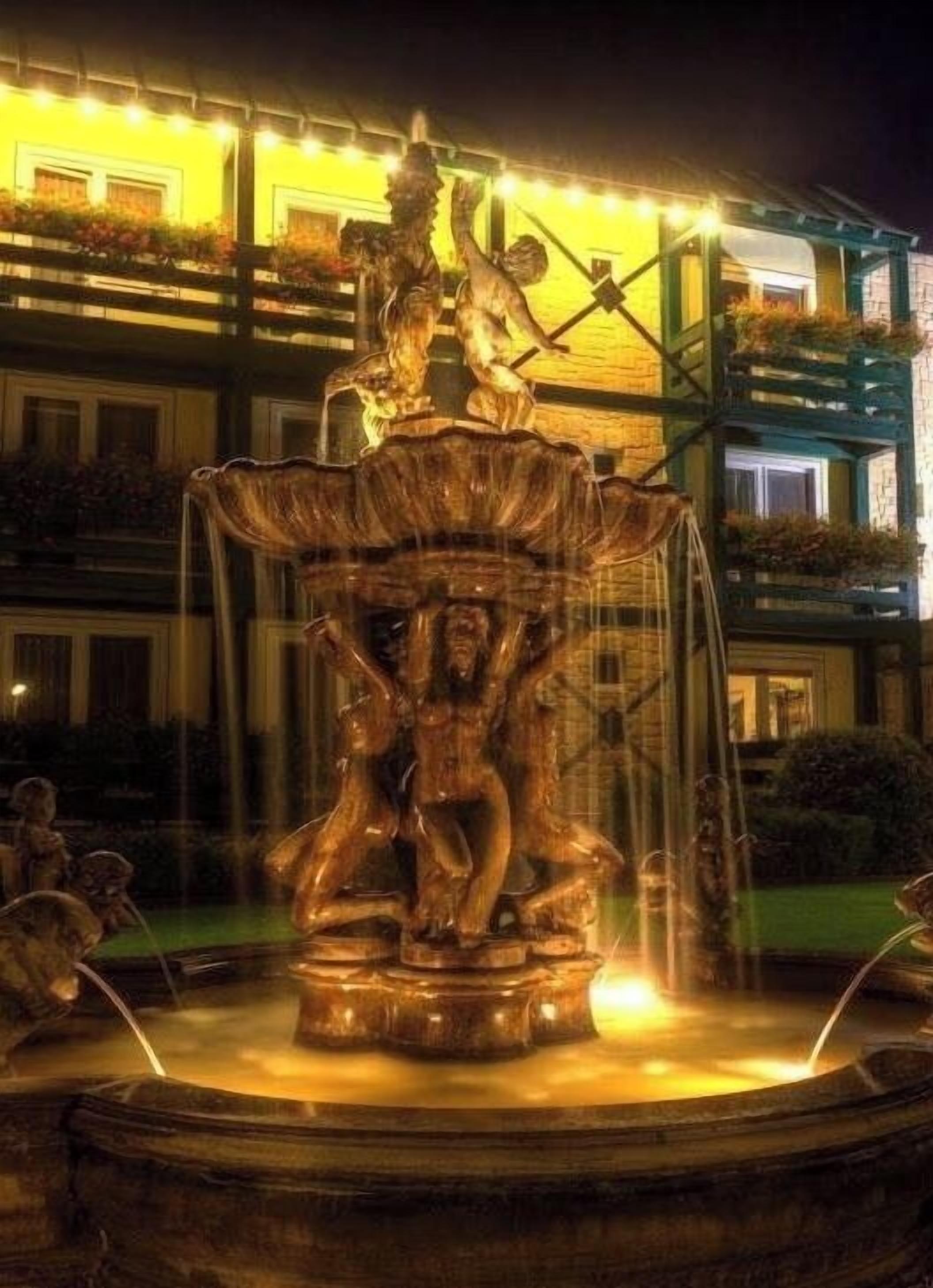 A fountain with a statue of a figure holding a staff, surrounded by water jets, set against a building with balconies and string lights.