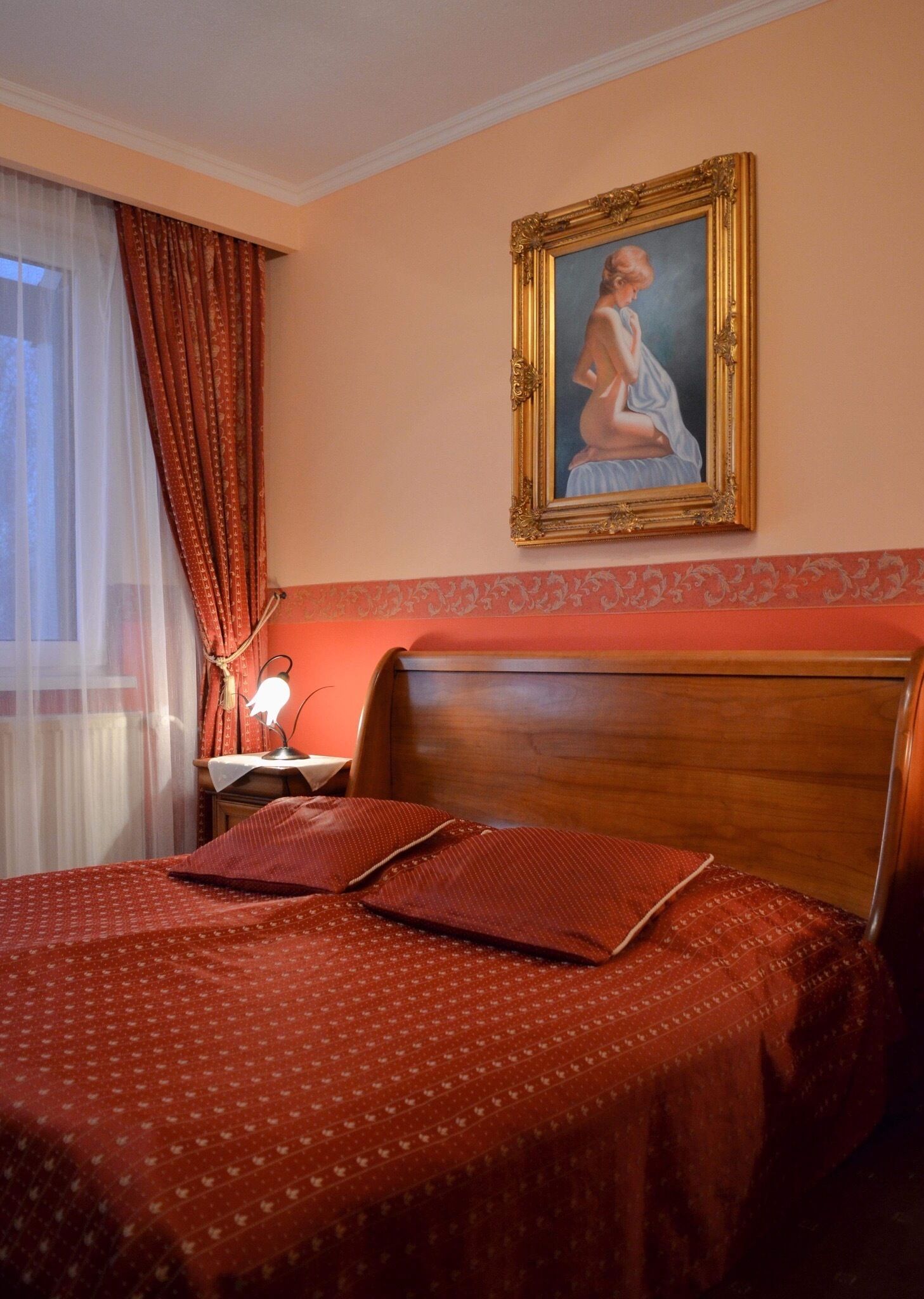 Habitación de hotel con una cama, una mesita de noche con lámpara, un cuadro en la pared y una ventana con cortinas.